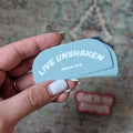 Hand holding a blue card with 'Live Unshaken' text against a patterned rug background