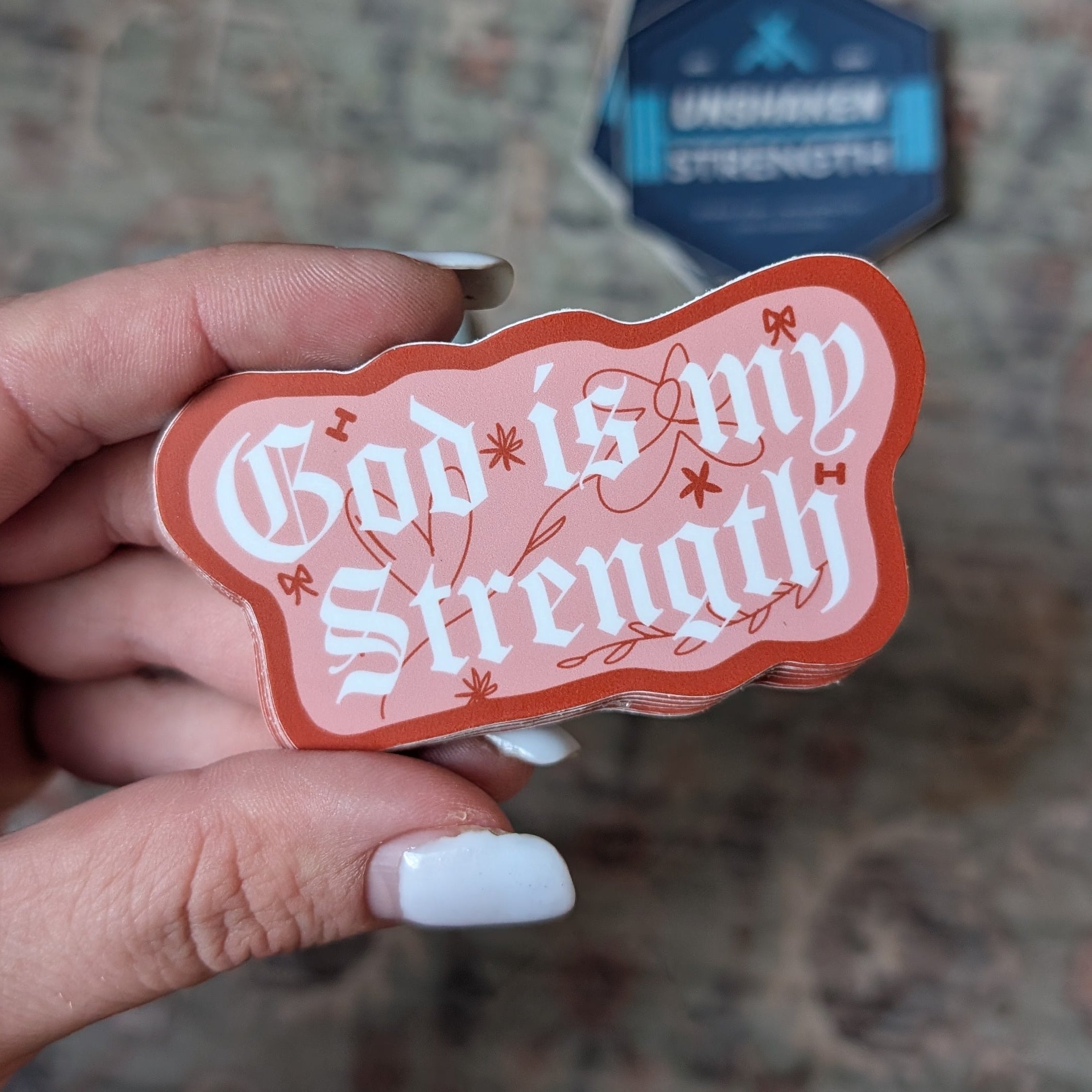 Hand holding a red and white sticker with 'God is my strength' text against a patterned background.