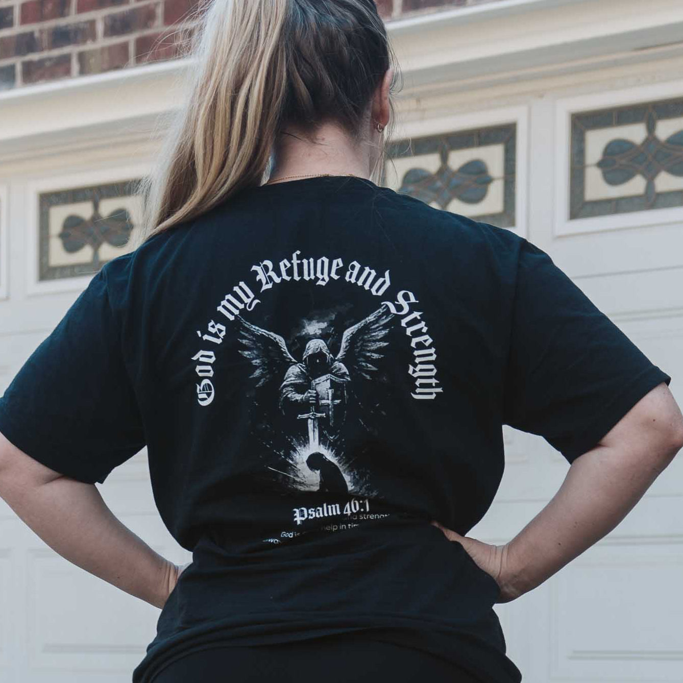 Person wearing a black t-shirt with a graphic design, standing in front of a garage.