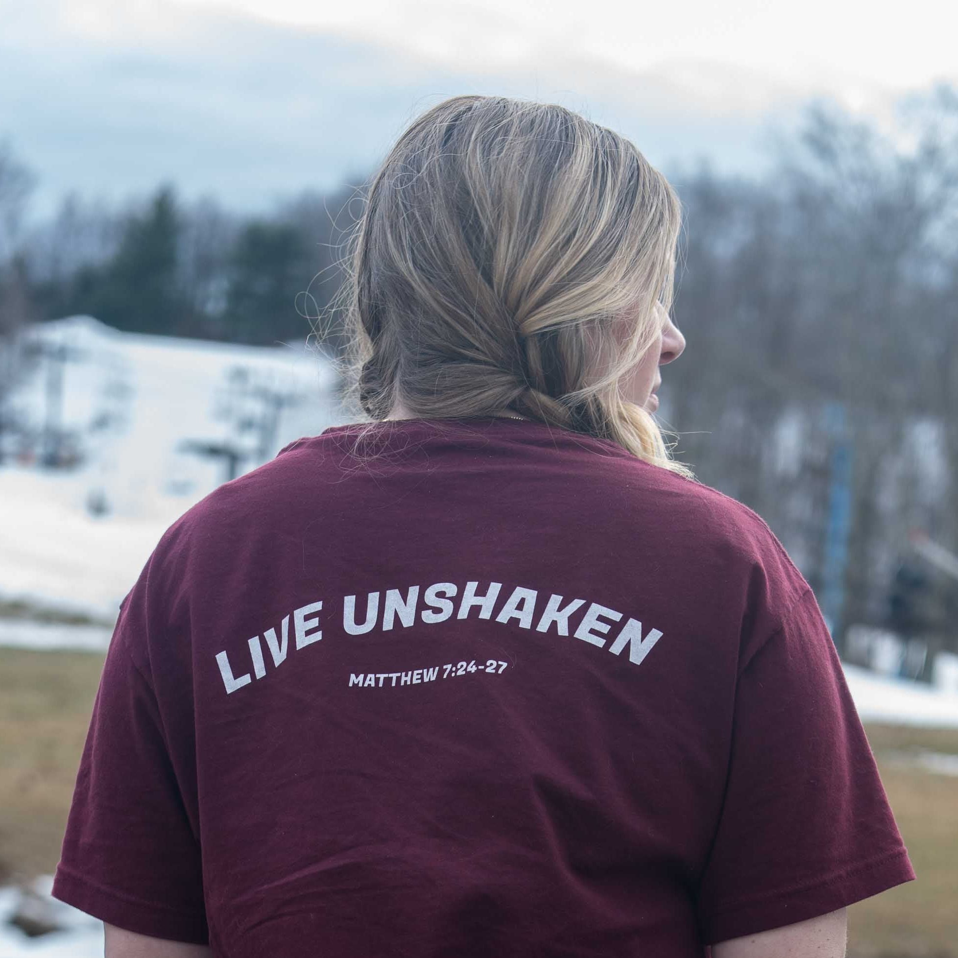 Person wearing a maroon t-shirt with 'LIVE UNSHAKEN' text outdoors.