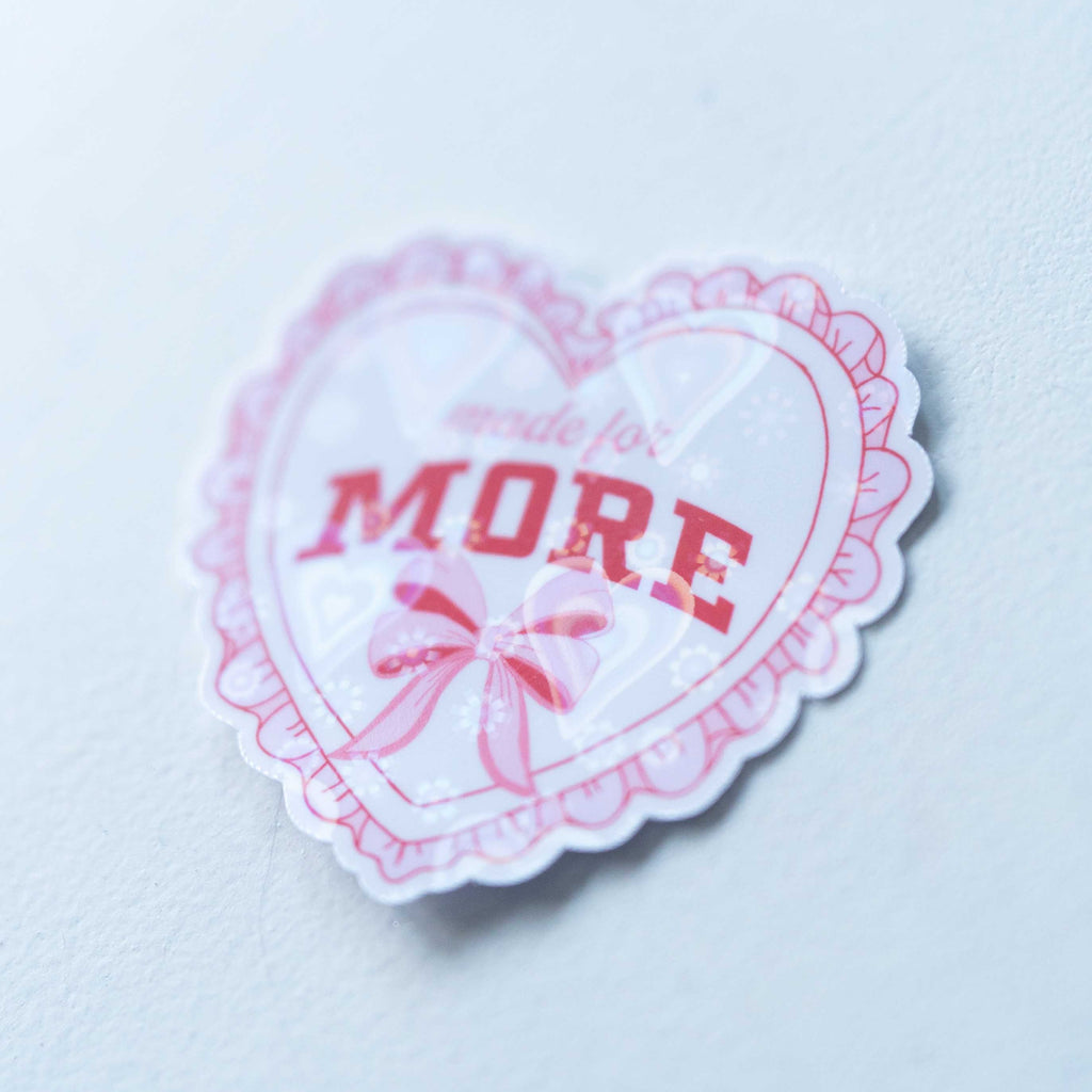 Heart-shaped sticker with 'MORE' text and a bow on a light blue background