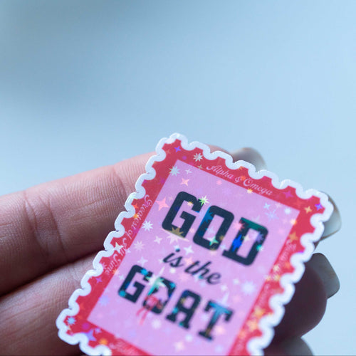 Hand holding a pink and black 'GOD is the GOAT' sticker against a blurred background