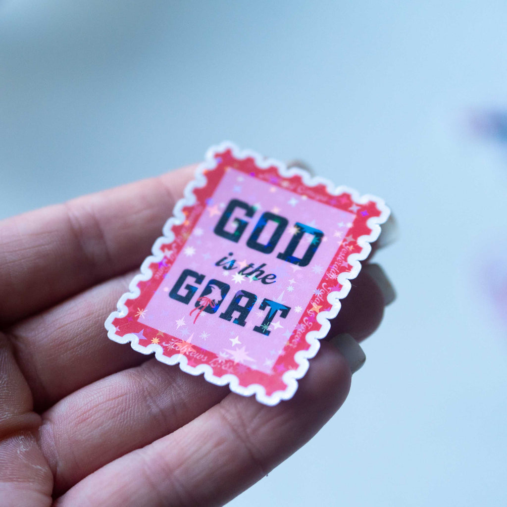 Hand holding a pink sticker with 'GOD is the GOAT' text against a blurred background